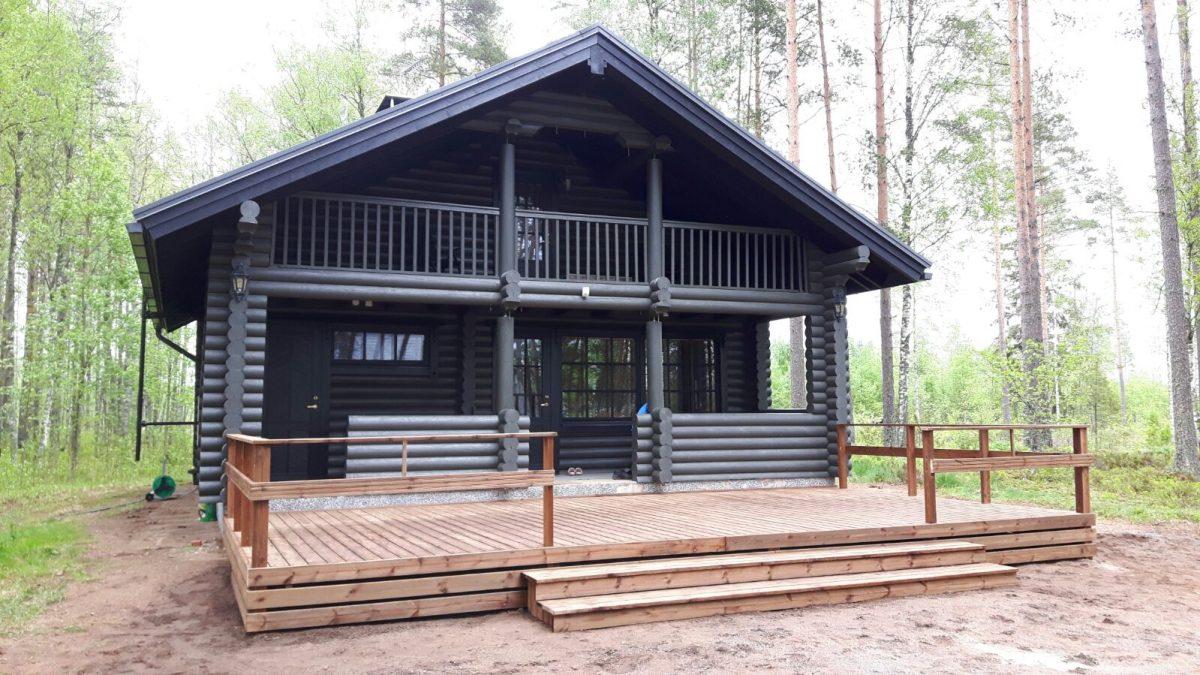 Tummanharmaa, kaksikerroksinen hirsimökki. Edessä iso puinen terassi. Yläkerrassa koko mökin levyinen parveke.