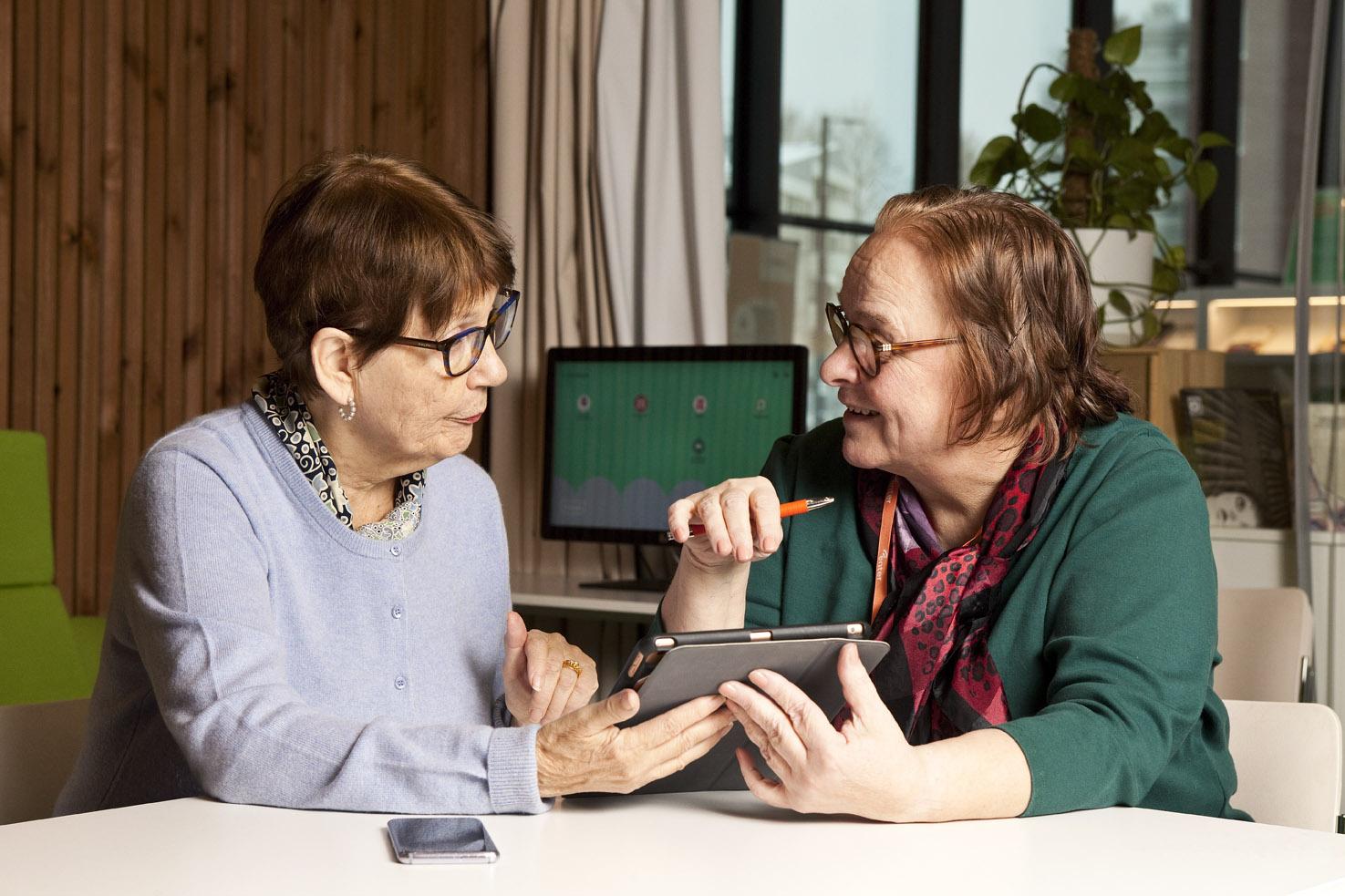 Kaksi seniorirouvaa keskustelee pöydän ääressä pidellen tablettia.