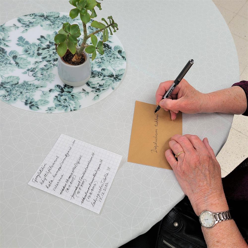 Kuvassa näkyvä käsi kirjoittaa kirjekuoreen "Digitaalinen tahtoni". Pöydällä on lista salasanoja.