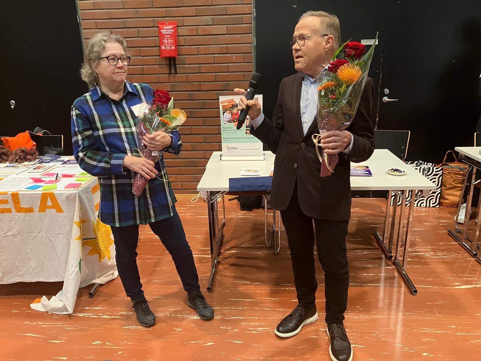 Jukka Tamminen puhuu mikrofoniin. Hänellä ja Ritva Lairalalla kukkakimput.