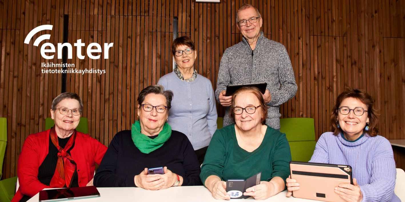 Seniorit ovat koolla pöydän ääressä ja katsovat kameraan hymyillen.