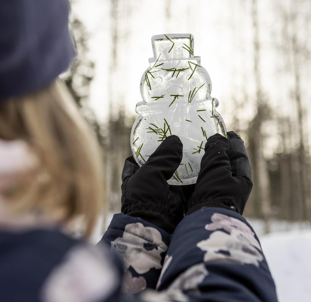 Toppavaatteisiin pukeutunut lapsi pitelee jäästä tehtyä lumiukkoveistosta. Veistokseen on tarttunut vihreitä havunneulasia. Taustalla näkyy talvinen metsämaisema.