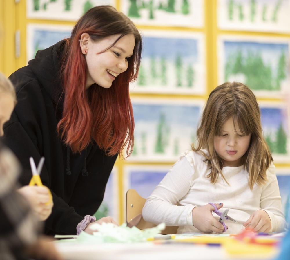 Kaksi lasta ja kaksi ohjaajaa istuu pyöreän matalan pöydän ympärillä. Toinen lapsista esittelee pöydällä olevaa taideteosta. Toisen ohjaajan kainalossa istuu musta pörröinen koira.