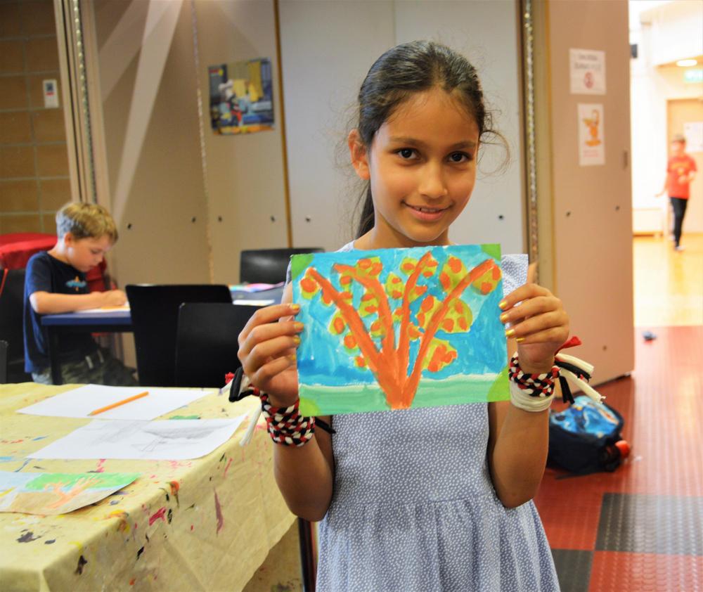 Lapsi esittelee kameralle maalaustaan. Taustalla toinen lapsi piirtää pöydän ääressä.