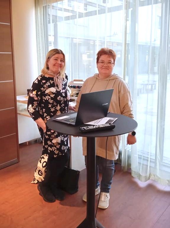Tarja Tachi ja Jaana Kesänen seisovat hymyillen pöydän vieressä jolla on tietokone