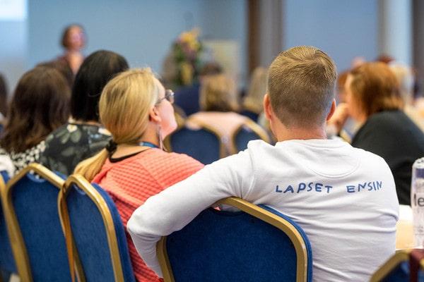 Kuvassa näkyy ihmisiä kokoustamassa isossa tilassa, etualalla on mies ja nainen selän puolelta kuvattuna. Toisen paidassa lukee lapset ensin, joka on MLL:n yksi sloganeista.