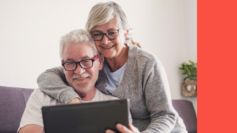Iäkäs nainen, jolla on lyhyet harmaat hiukset, istuu terassilla ja hymyilee kameralle. Hän pitelee kahvikupista, joka on pöydällä hänen edessään.