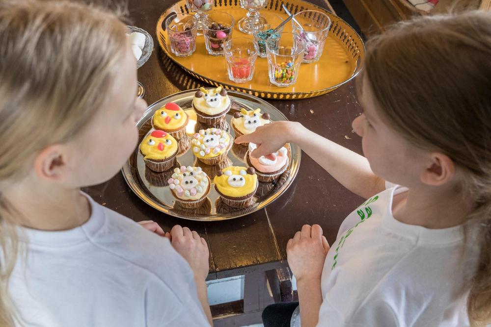 Kokkailua Märynummella -kerhon kuvassa on kaksi tyttöä pöydän ääressä. Heidät on kuvattu takaa päin yläviistosta. Pöydällä on värikkäästi koristeltuja kuppikakkuja, joista tulee mieleen pääsiäinen ja joita tytöt katselevat.