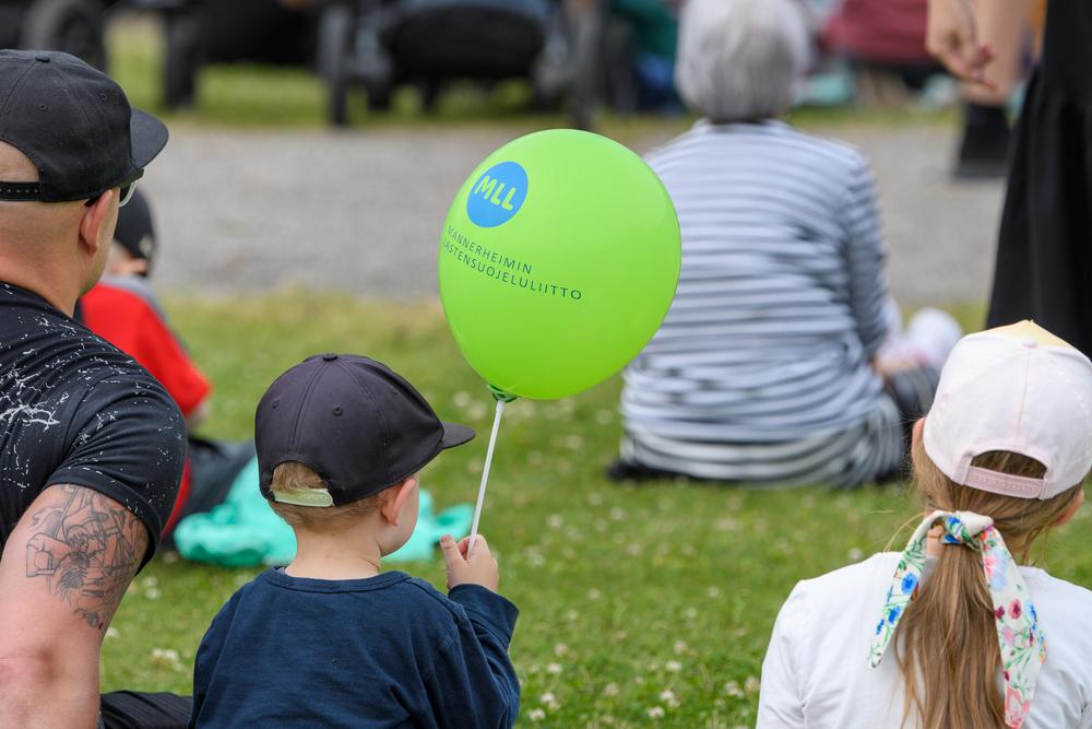 Lapsi istuu puistossa aikuisen seurassa ja pitää kädessään vihreää Mannerheimin lastensuojeluliiton ilmapalloa. Ympärillä on muita perheitä.