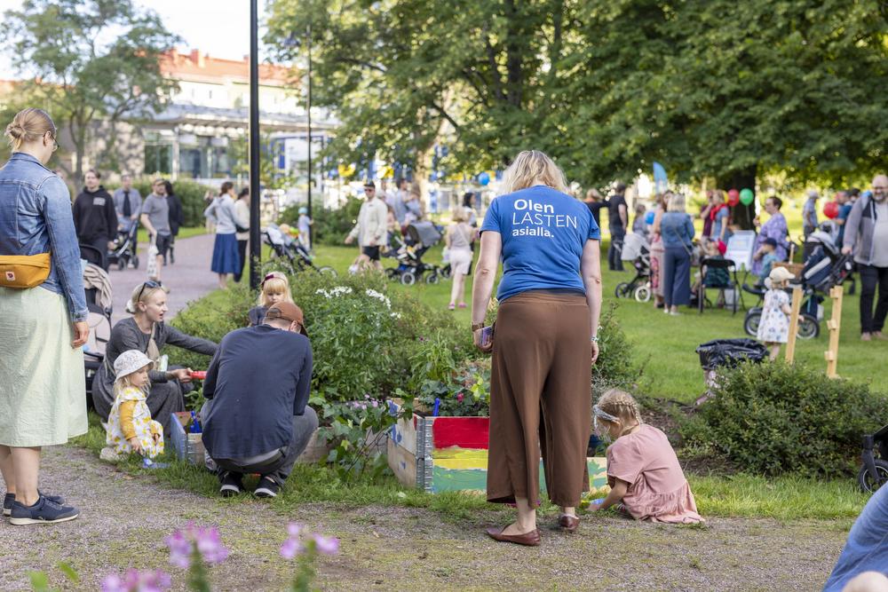 Puistotapahtuma, jossa on lapsiperheitä. MLL:n vapaaehtoinen tarkastelee istutuslaatikkoa, jonka äärellä istuu lapsi. Vapaaehtoisella on sininen t-paita, jossa lukee "Olen lasten asialla".