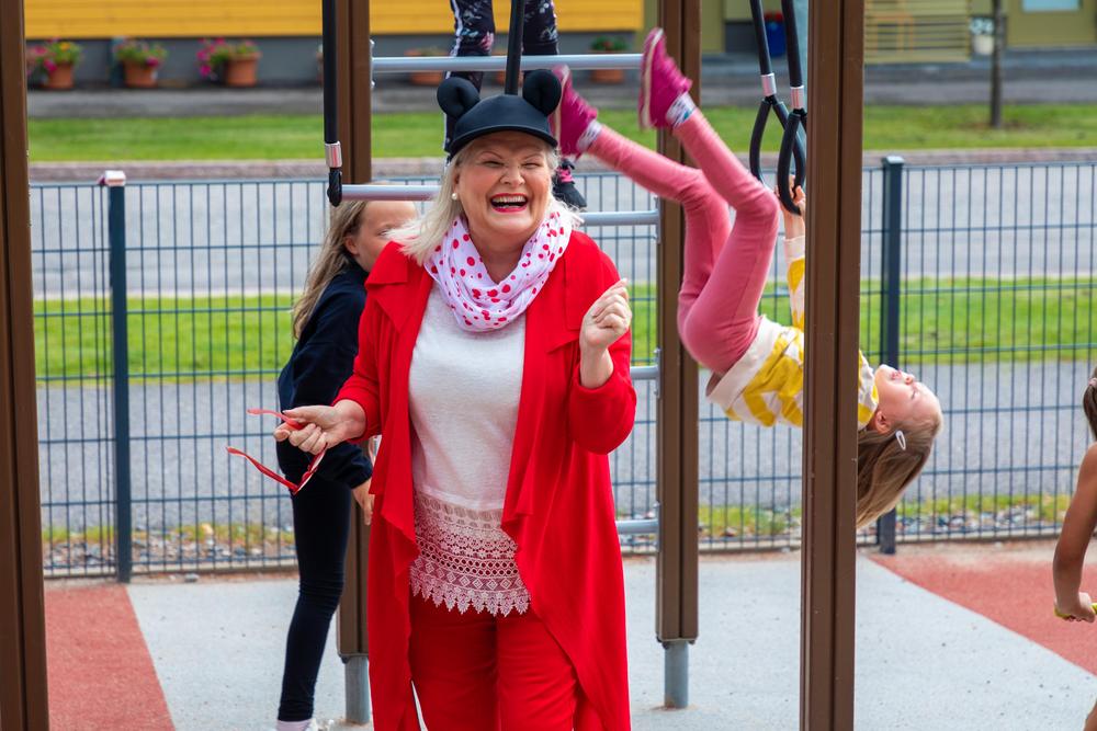 Mikki Hiiri -hattuun pukeutunut nainen nauraa leikkipuistossa. Lapset temppuilevat kiipeilytelineessä.