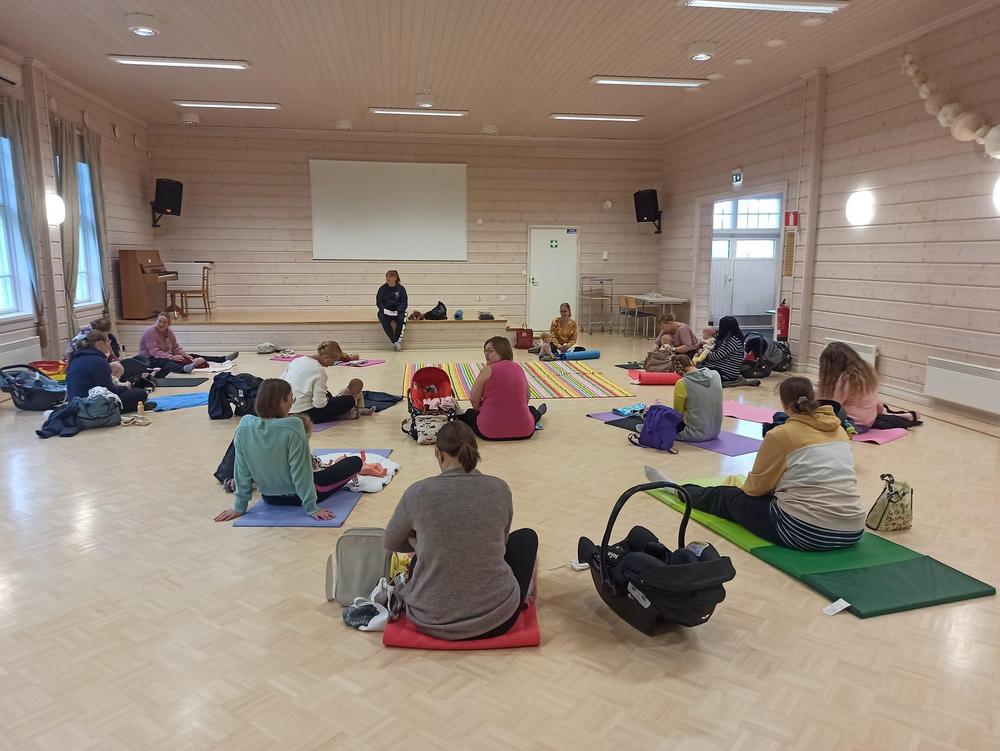 Kuvan takaosassa vauvahieronnan vetäjä odottamassa ohjauksen alkua. Hänen edessään on puoliympyrään asettautuneena äitejä ja vauvoja jumppamattoineen valmistautumassa.