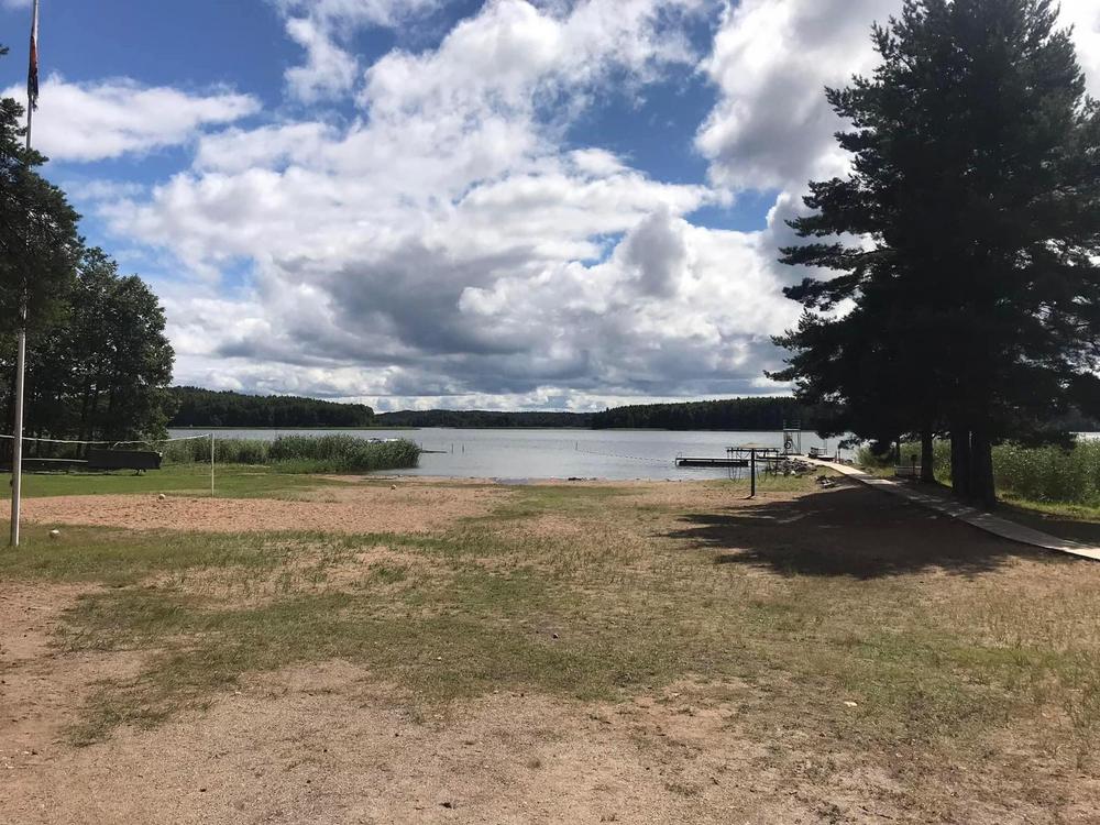 Oikeassa reunassa kulkee puinen laituri hiekkarannan läpi. Laituri jatkuu merelle asti, ja loppupäässä näkyy hyppytorni ja uimatikkaat. Keskellä näkyy tyhjää uimarantaa ja vasemmalla lentopalloverkko. Taka-alalla siintää meri.