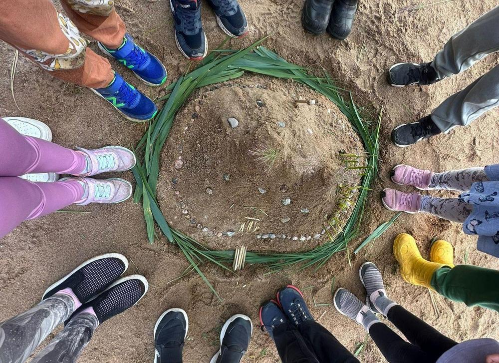 Keskellä ylhäältä kuvattu kaisloilla ja muilla luonnonmateriaaleilla koristeltu hiekkalinna. Linnan ympärillä seisoo lapsia, joista näkyvät vain jalat.