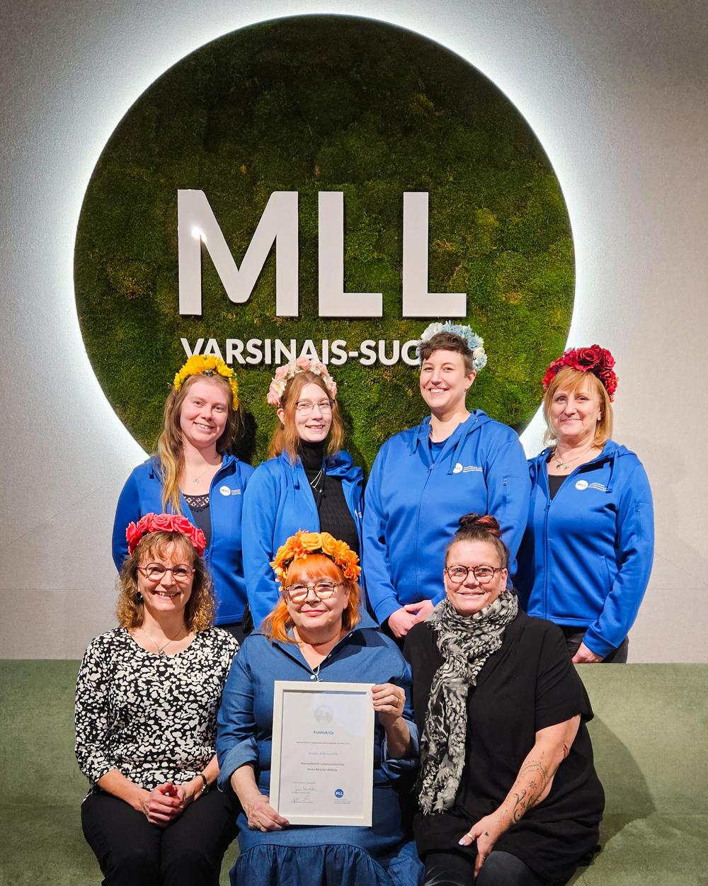 Etualalla istuu kolme ja takana seisoo neljä naista. Etualan keskimmäisellä naisella on Vuoden yhdistys -kunniakirja käsissään. Kaikki katsovat hymyillen kameraan ja taustalla näkyy iso MLL Varsinais-Suomi-logo.