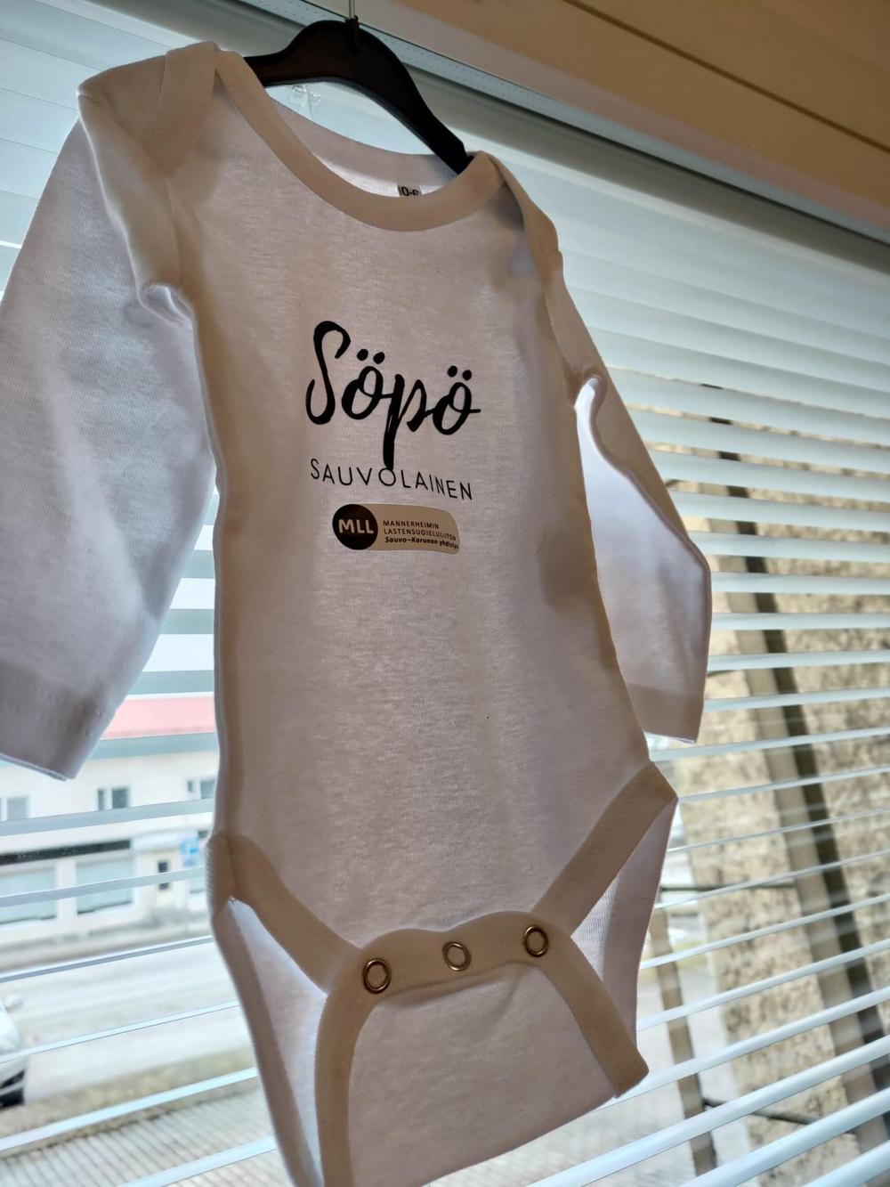 Kuvassa on valkoinen vauvanbody, jossa rinnassa on kaunokirjoituksella teksti: "Söpö sauvolainen" ja sen alla MLL Sauvo-Karunan yhdistyksen logo.