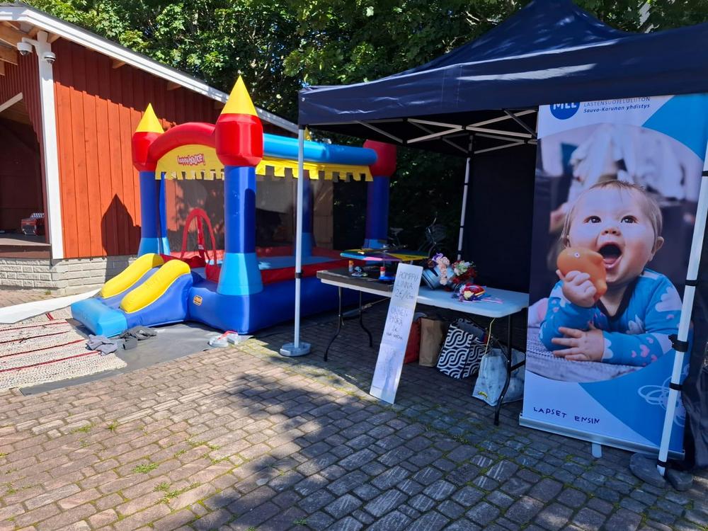Vasemmalla pomppulinna, jonka vieressä telttakatoksen alla pöydällä onnenpyörä ja kukkapantoja. Oikeassa reunassa MLL:n ständi.