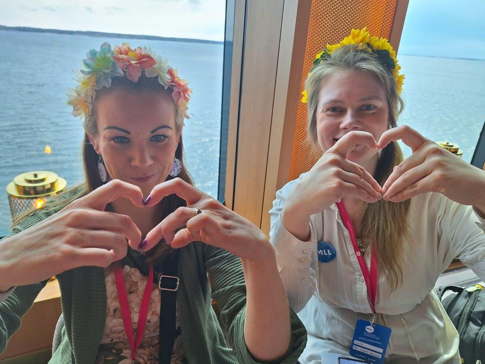 Kaksi naistaa istuu risteilyllä laivan ikkunalaudalla kukkapannat päässään ja näyttävät sormillaan sydäntä.