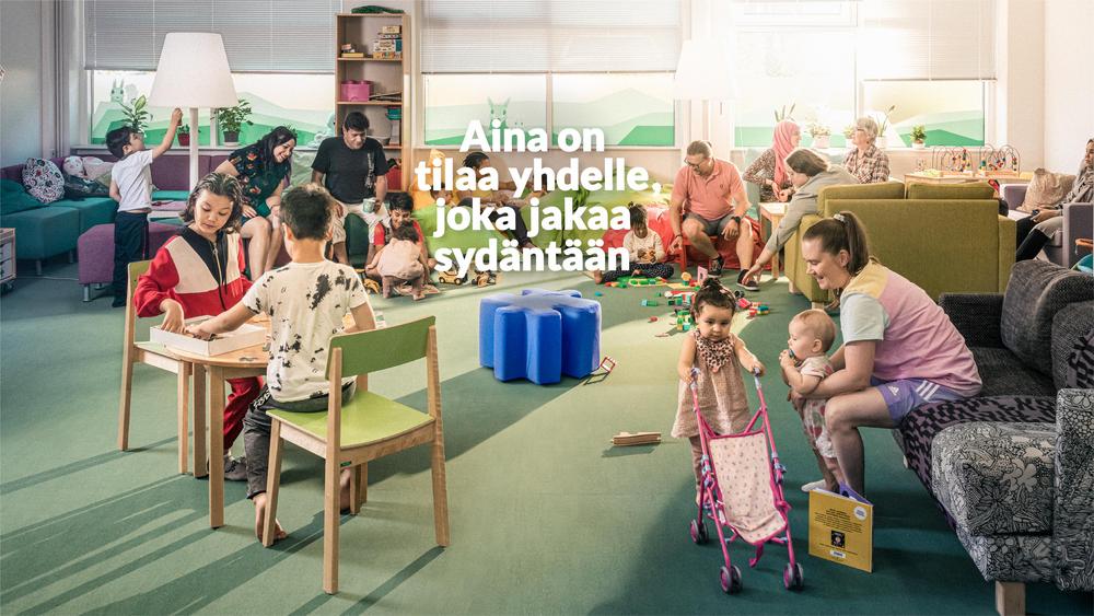 Lapsia vanhempineen ja isovanhempineen leikkimässä perhekahvilassa. Keskellä sininen istuin tyhjänä.
