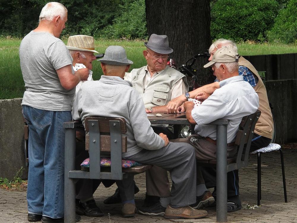 Kuvassa on vanhempia miehiä pelaamassa korttia ulona pöydän ääressä. Taustalla viheriöi nurmikko. 