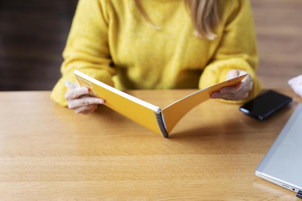 Keltapuseroinen nainen lukee kierrevihkosta muistiinpanoja Palvelualojen ammattiliiton PAMin sopimasta kaupan esihenkilöiden työehtosopimuksesta. Sopimus koskee työaikalain alaisia vähittäiskaupan esihenkilöitä sekä Kaupan  työehtosopimuksessa tarkoitettujen pika- ja noutotukkujen esihenkilöitä.