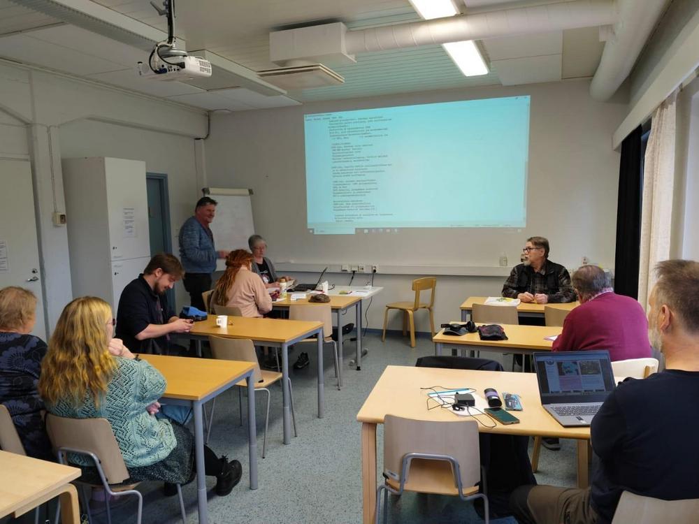 Luento tai kokous Sepänkeskuksen Koeputki-tilassa. Ihmiset istuvat pöytien ääressä kuin koulun luokassa. Esittäjä näyttää edestä tekstiä videotykiltä.