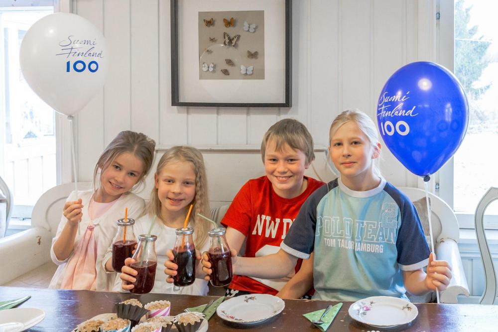 Kuvassa istuu neljä lasta ruokapöydän ääressä. Kaikilla lapsilla on smoothiet kädessä ja kahdella lapsella on toisessa kädessä ilmapallot. 