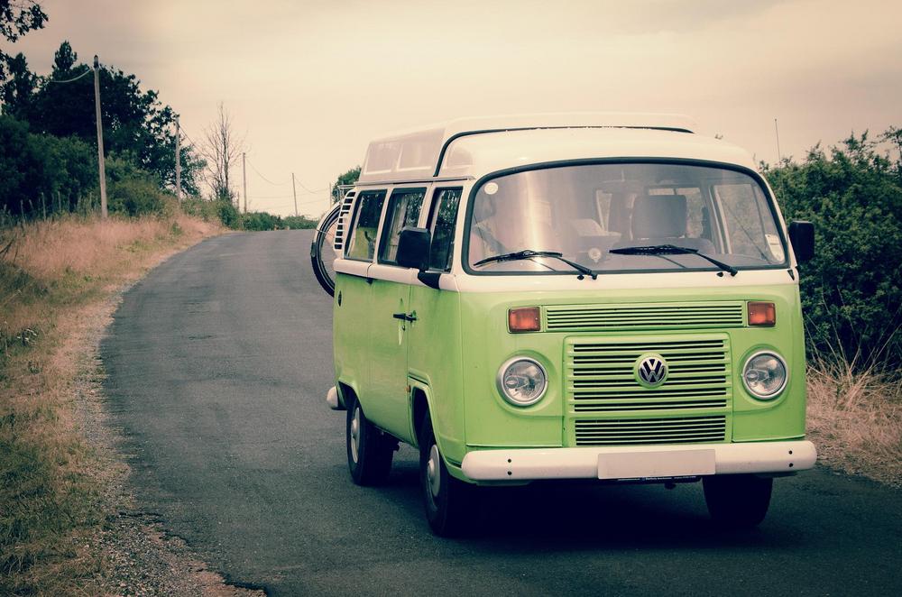 Kuvassa vanha vihreä-valkoinen Volkswagen -bussi asfaltoidulla tiellä, takana pensaikkoa.