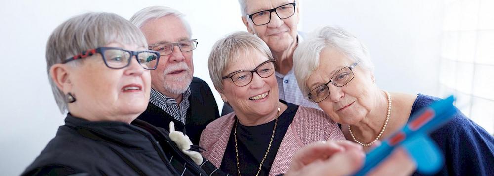 Kuvassa harmaapäisiä senioreita, edessä kolme rouvaa, heidän takanaan kaksi herra tutustuvat senioriliiton App'n lataamiseen matkapuhelimelle.