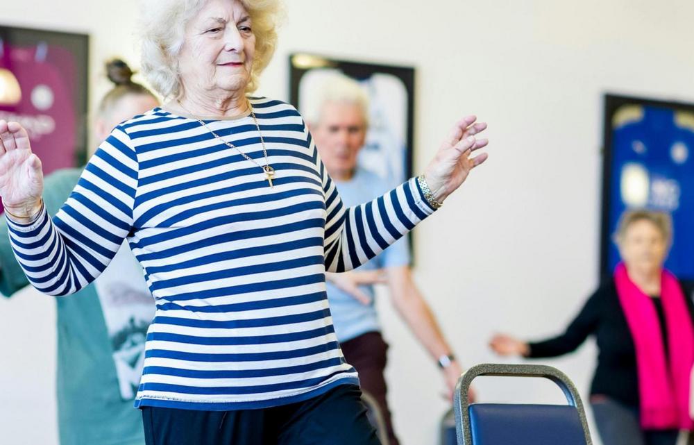 Ikäihmisiä tuolijumpassa, etualalla nainen sini-valkoraitainen paita päällä, takana rouva, jolla on punainen huivi kaulassa.