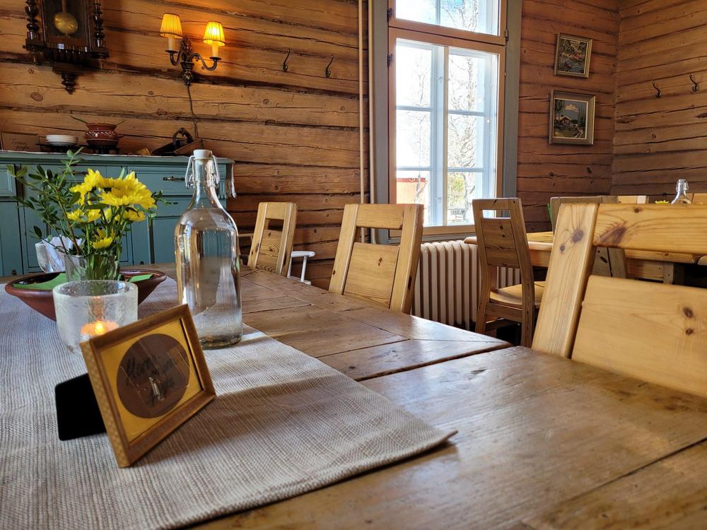 Maalaistalon hirsiseinäinen ruokailutila, pöydällä vanha pullo ja keltainen kukka.