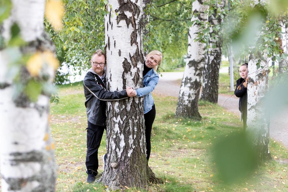 Valokuva: Kuva puistokoivikosta. Etualalla olevan ison koivun takaa kurkistaa toiselta puolelta mies, toiselta nainen. He pitävät toisiaan kädestä puun etupuolelta. Taempana olevan puun takaa kurkistaa teini-ikäinen tyttö.