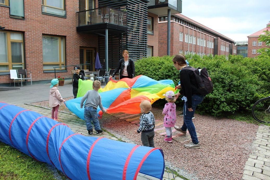 Valokuva: Kesäinen päivä ulkona. Joukko aikuisia ja lapsia pitää välissään värikästä leikkivarjokangasta ja heiluttaa sitä ilmassa. 