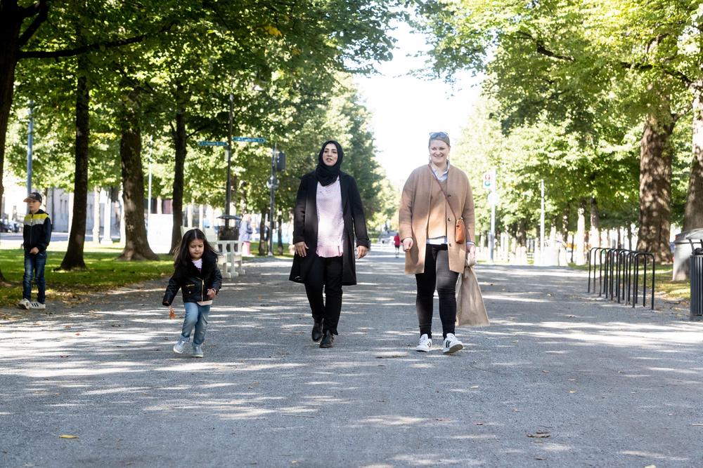 Valokuva: Aurinkoinen Hämeenpuisto, keskellä leveä hiekkatie, jota puut reunustavat. Keskellä kävelee kaksi naista rinnakkain, toisella on hijab. Vieressä kulkee leikki-ikäinen lapsi.