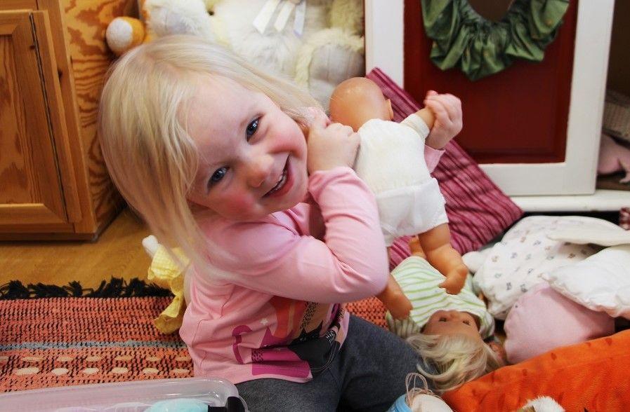 Valokuva: Kuva on lähikuva nukkea nukkeaan puristavasta ja suoraan kameraan leveästi hmyillen katsovasta lapsesta.