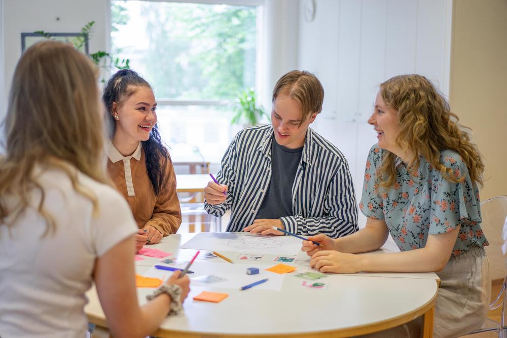 Valokuva: Nuoret istuvat pöydän ääressä nauraen, pöydällä on paljon koulutavaroita.