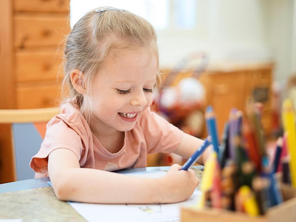 Valokuva: Kuvassa on lapsi värittämässä värityskuvaa pöydän ääressä. Etualalla erivärisiä puukyniä telineessä.