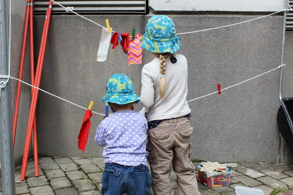 Valokuva: Kaksi lasta, isompi ja pienempi, seisovat harmaan rakennuksen edessä ollen selin kameraan. Heidän eteensä on pingotettu kaksi eri korkeuksilla olevaa pyykkinarua. Pyykkinarussa on kiinni värikkäitä kangastilkkuja. 