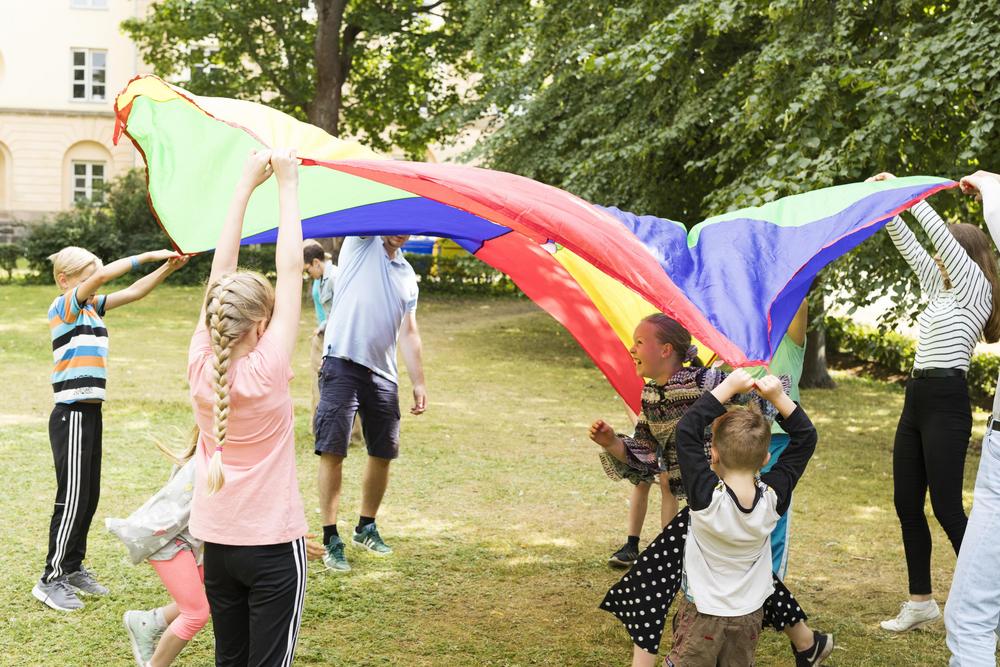 Lapset ja aikuiset leikkivät leikkivarjon alla nurmikentällä.