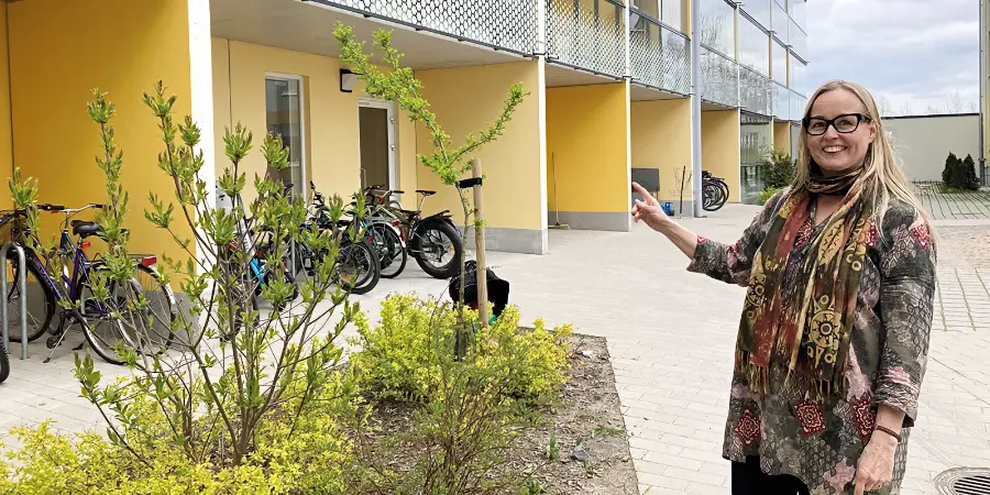 Hymyilevä vaaleahiuksinen nainen osoittaa kerrostalon pihalla olevaa ruukkuistutusta. 