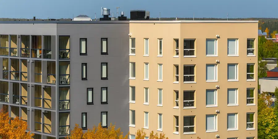 Vierekkäin sijaitsevat vaalea ja tummanharmaa kerrostalo syksyn väreissä olevien lehtipuiden ympäröimänä kaupunkimaisemassa.