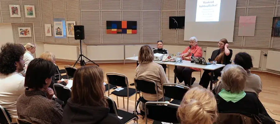 Tapahtumatilan esiintyjien pöydän äärellä istuu kolme naista kasvot kohti edessä istuvaa yleisöä. Yksi esiintyjistä puhuu juuri mikrofoniin.