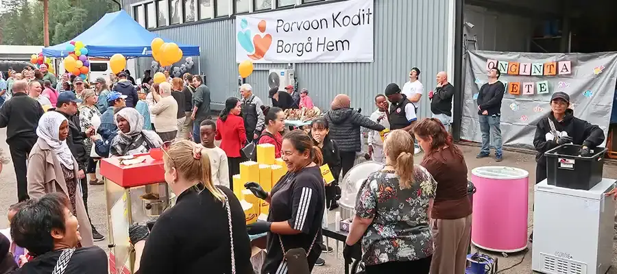 Harmaan hallin edustalla ulkona on suuri ryhmä ihmisiä. Tarjolla on ruokaa sekä ilmapalloja ja hallin ulkoseinälle on ripustettu teksti "Porvoon Kodit, Borgå Hem".