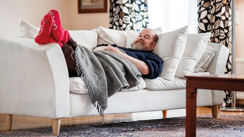 Parrakas mies lepäämässä valkoisella sohvalla viltin alla punaiset villasukat jalassaan.
