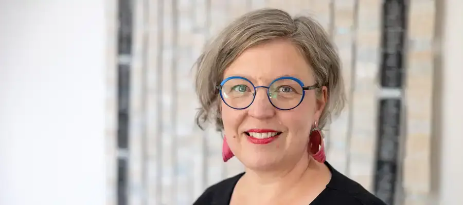 Kasvokuvassa hymyilevä silmälaseja sekä punaisia riippuvia korvakoruja käyttävä nainen.