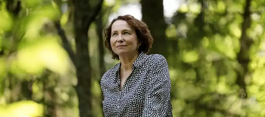 Ruskeahiuksinen nainen seisoo hymyilemässä vehreitä lehtipuita taustallaan.