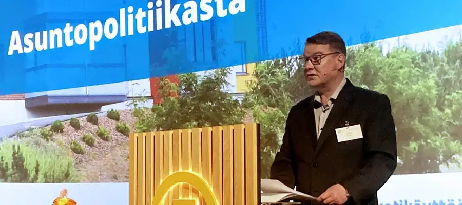 Tummaa pukua ja kauluspaitaa sekä silmälaseja käyttävä mies esiintymislavalla pitämässä puhettaan.