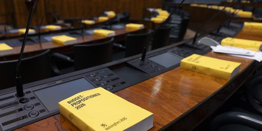 Eduskunnan istuntosalin pöydillä on keltaisia vuoden 2026 talousarviokirjoja.