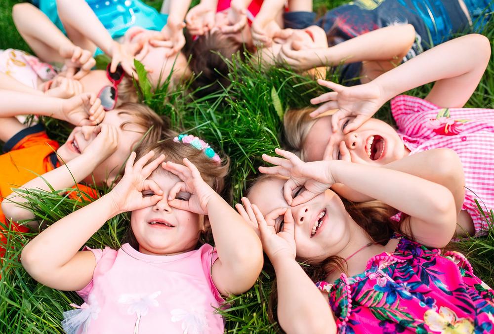 Lapset makaavat nurmikolla selällään ja ympyrässä kurkistellen peukalolla ja etusormella tehdylillä kiikareille. Kuva Adobe Stock.