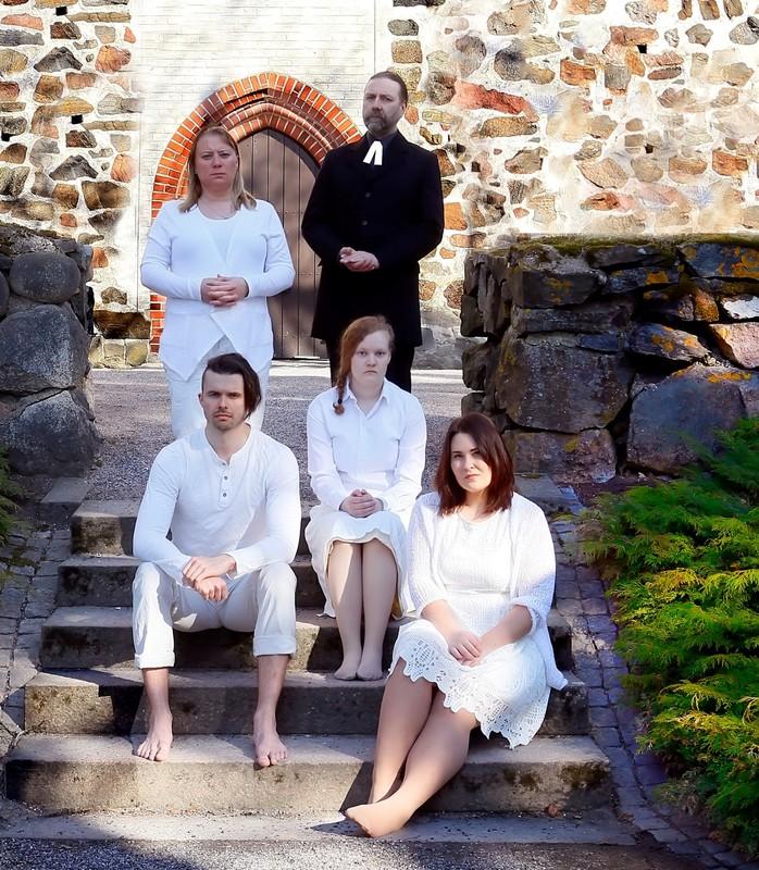 Papin perheen näyttelijät roolivaatteissa kirkon portailla. Etualalla istuvat lapset valkoisissa vaatteissa. Takana seisovat Elizabeth valkoisessa vaatetuksessa sekä Pastori mustassa asussaan liperit kaulassa.